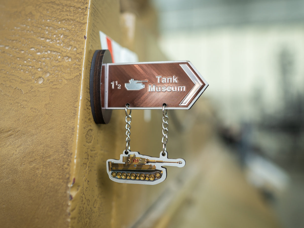 A small metal sign pointing to the Tank Museum, mounted on a gold-toned surface with two hanging tank-shaped charms below.