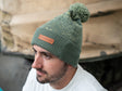 A man modelling a two-tone green knitted design bobble hat, with The Tank Museum logo on the cuff. Finished with a debossed faux-leather Tank Museum tab.