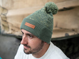 A man modelling a two-tone green knitted design bobble hat, with The Tank Museum logo on the cuff. Finished with a debossed faux-leather Tank Museum tab.