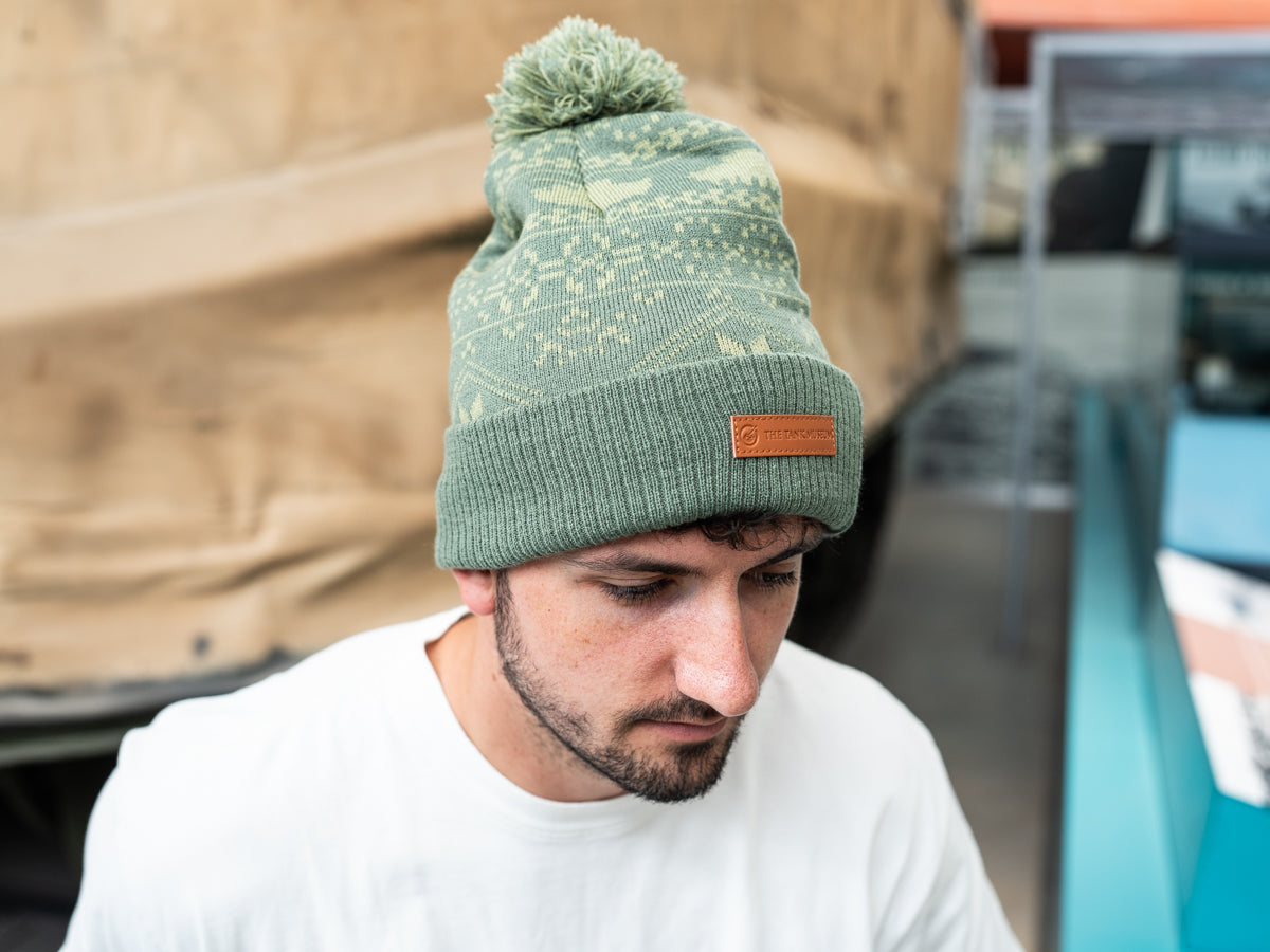A man modelling a two-tone green knitted design bobble hat, with The Tank Museum logo on the cuff. Finished with a debossed faux-leather Tank Museum tab.