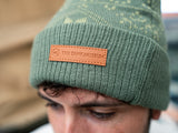 A man modelling a two-tone green knitted design bobble hat, with The Tank Museum logo on the cuff. Finished with a debossed faux-leather Tank Museum tab.