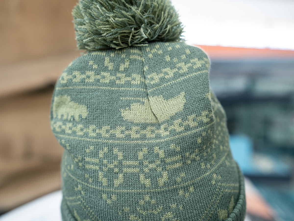 A man modelling a two-tone green knitted design bobble hat, with The Tank Museum logo on the cuff. Finished with a debossed faux-leather Tank Museum tab.
