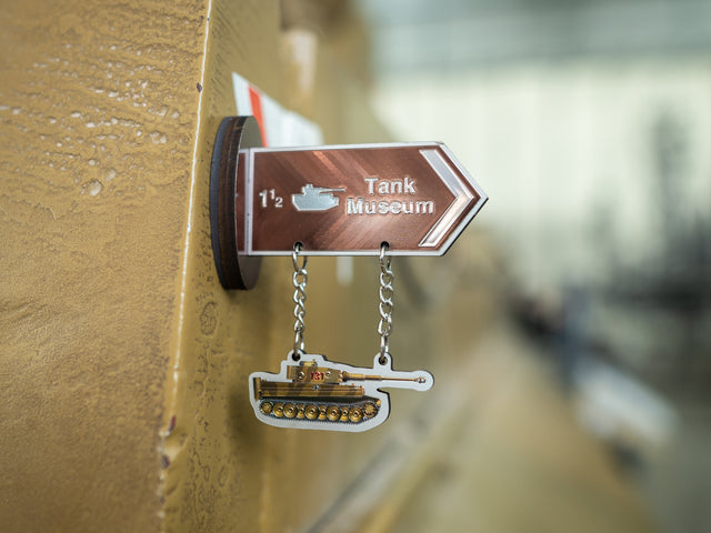 A small metal sign pointing to the Tank Museum, mounted on a gold-toned surface with two hanging tank-shaped charms below.